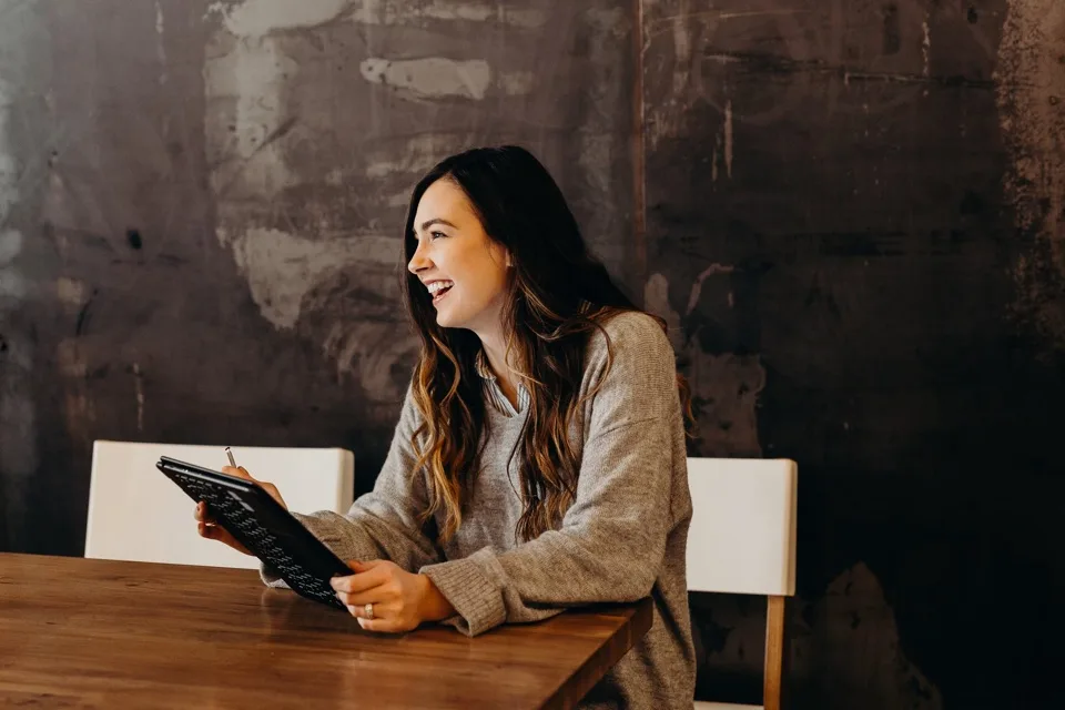 A smiling person holding a tablet at a wooden table.