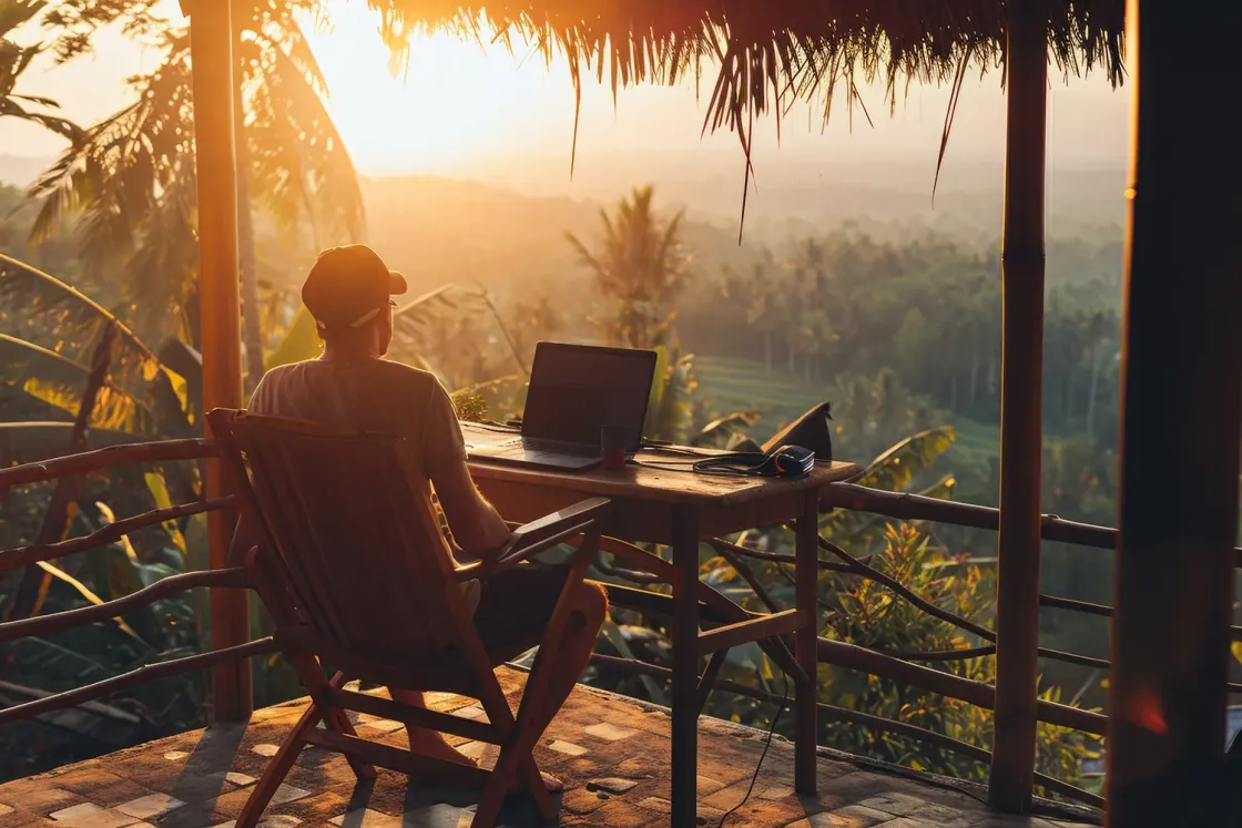 A remote worker with a laptop on a balcony above green hills at sunrise.